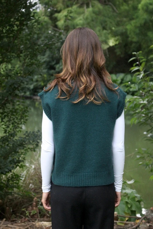 Green possum and merino wool vest on female model in park, back view. Lightweight, warm and comfortable women's wool winter vest. Made in New Zealand