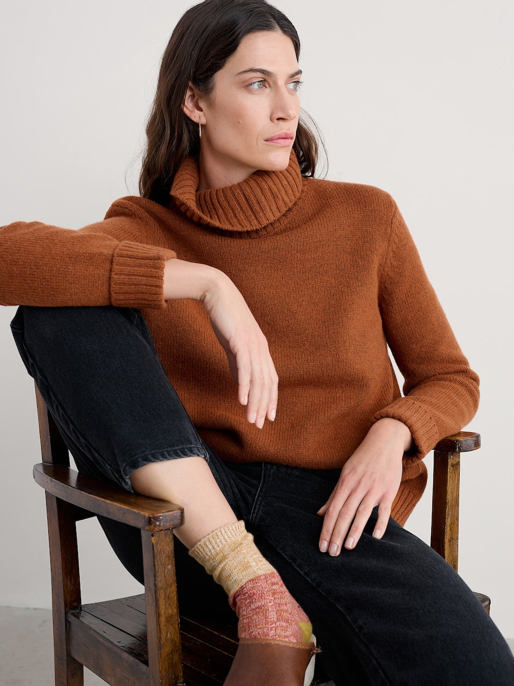 Woman wearing a brown sweater sitting on a wooden chair against a plain background