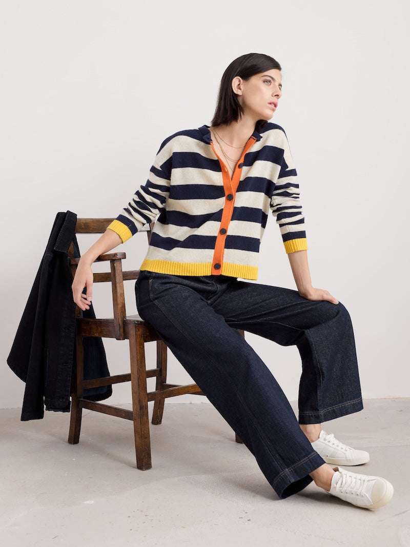 Woman sitting on a wooden chair wearing a striped sweater with orange buttons.