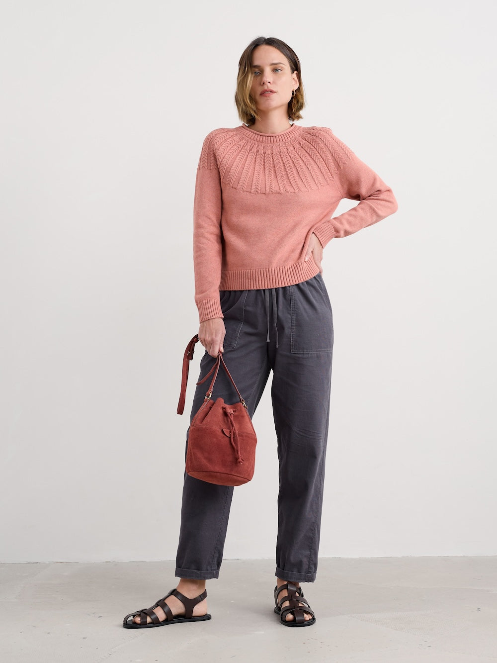 Woman wearing a pink sweater and dark pants, holding a brown bag against a white background