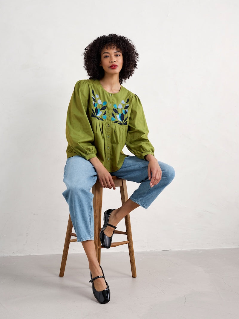 Woman sitting on a stool wearing a green embroidered blouse and light blue jeans against a white background