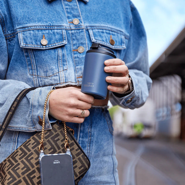 Denim blue Fressko Ceramic reusable coffee cup with black spill proof lid being held by woman. Stays hot for up to 3 hours. Chemical-free, lightweight & insulated stainless steel coffee cup