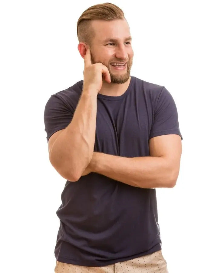 A smiling man with a beard and short haircut wearing a dark navy blue bamboo t-shirt and beige shorts, posing with his hand on his chin against a solid white background.