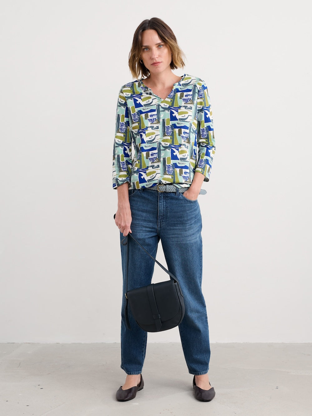 Woman wearing a patterned blouse and jeans, holding a black bag against a white background
