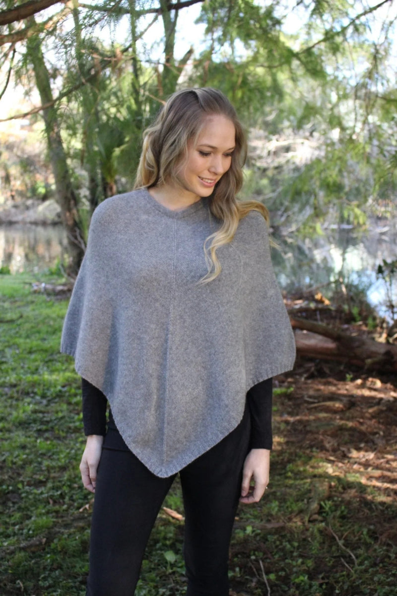 Silver grey possum and merino wool poncho on female model standing in park. Warm, soft, lightweight possum and merino wool. Lothlorian