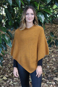 Gold possum and merino wool poncho on female model standing in garden. Warm, soft, lightweight possum and merino wool. Lothlorian