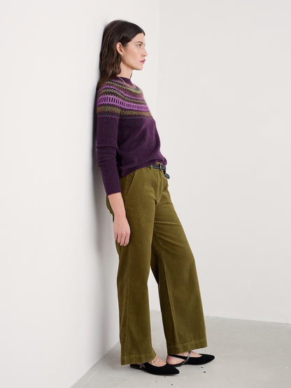 Woman wearing a purple sweater with a pattern and green pants against a white wall.