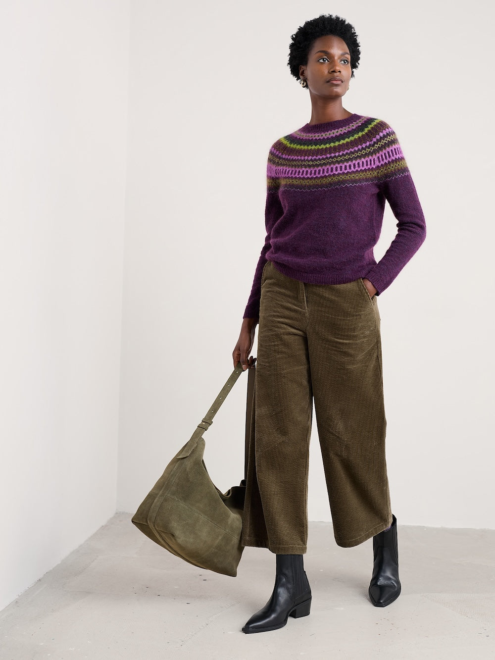 Woman wearing a purple sweater with a patterned yoke, holding a green bag, and standing against a white background.