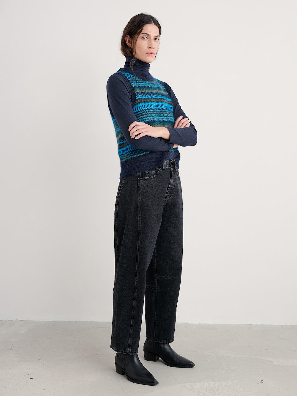 Person wearing a patterned turtleneck, vest and black pants on a plain background