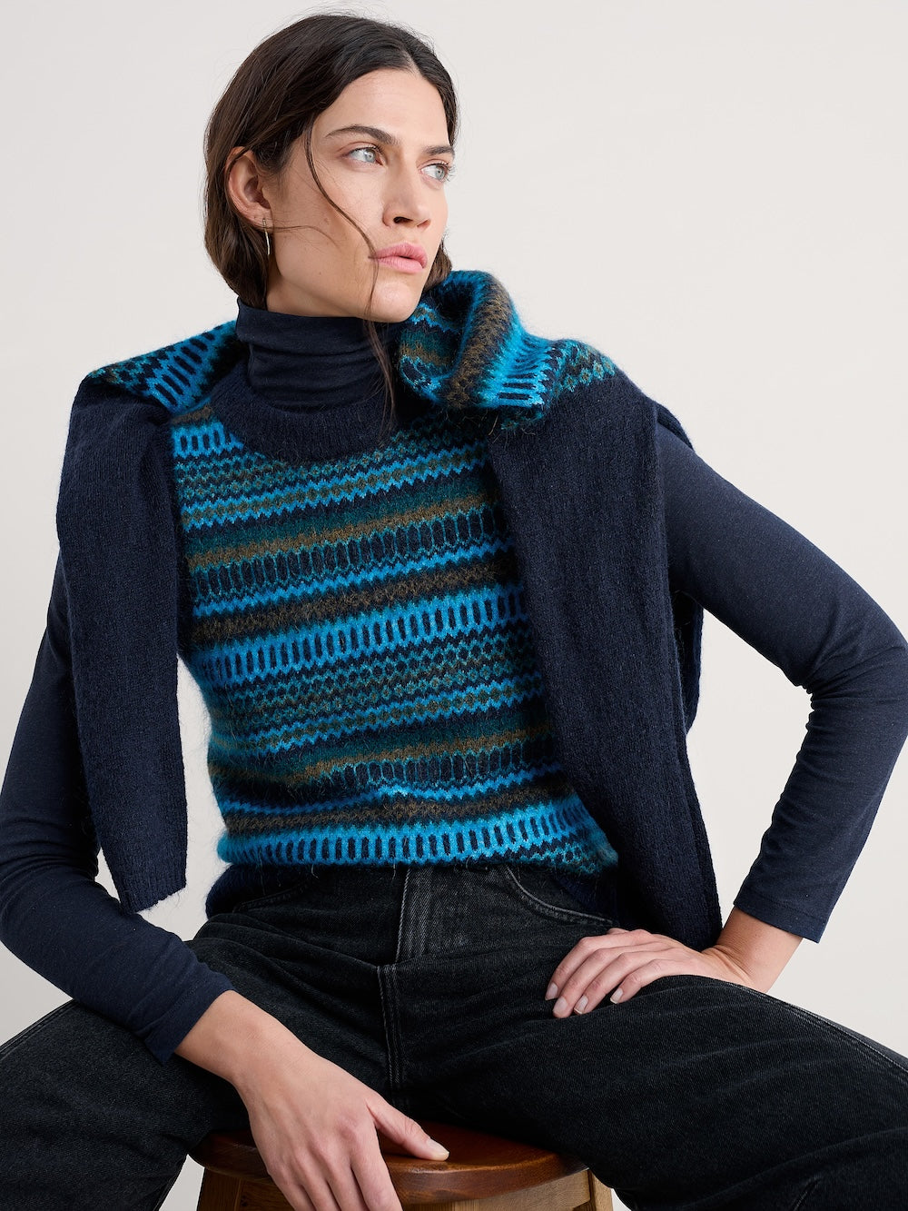 Woman wearing a blue and black striped vest over a dark shirt, sitting on a stool against a plain background.