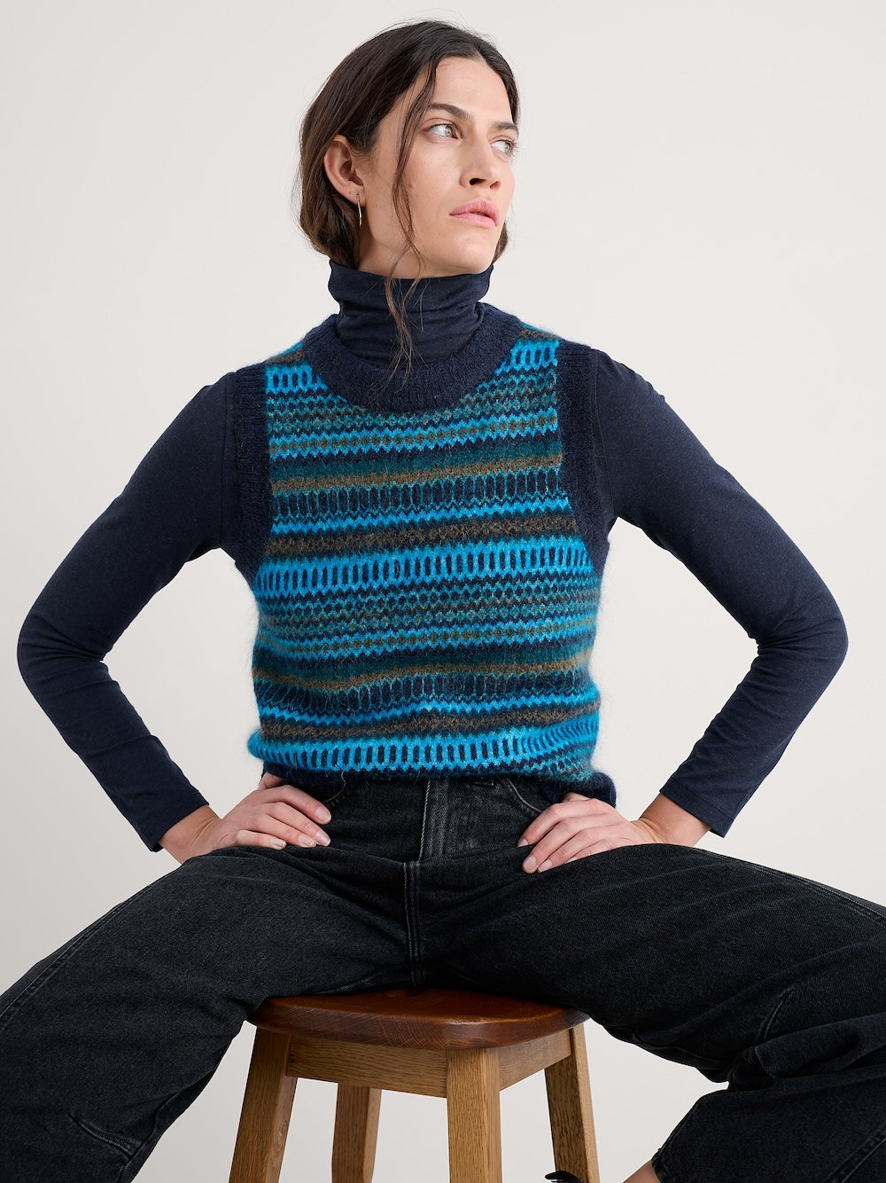 Person wearing a blue and black striped knit vest over a dark long-sleeve shirt, sitting on a wooden stool against a plain background.