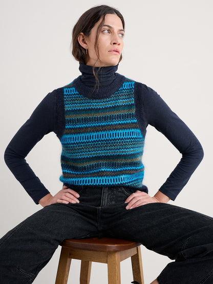 Person wearing a blue and black striped knit vest over a dark long-sleeve shirt, sitting on a wooden stool against a plain background.
