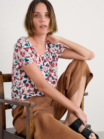 Woman sitting on a wooden chair wearing a colorful floral top and brown pants.