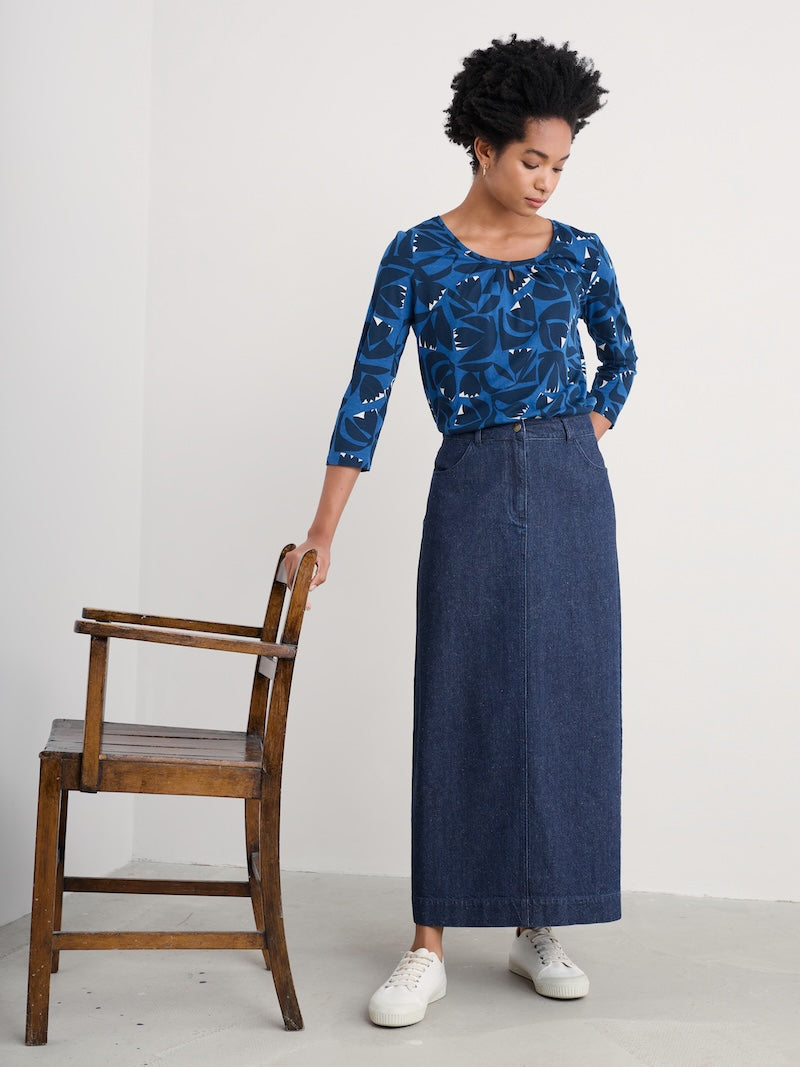 Woman wearing a blue patterned top and denim skirt standing next to a wooden chair against a white background