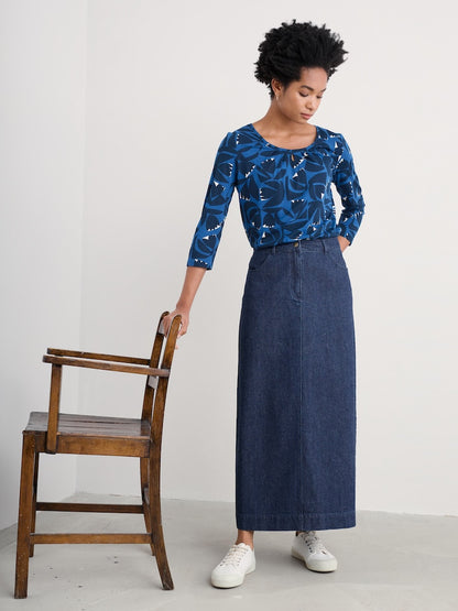 Woman wearing a blue patterned top and denim skirt standing next to a wooden chair against a white background