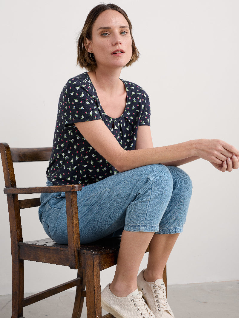 Woman sitting on a wooden chair wearing a navy patterned top and blue jeans.