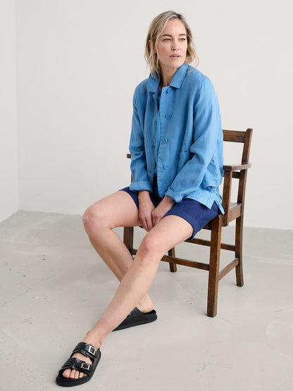 Woman wearing a blue shirt and shorts sitting on a wooden chair against a white wall.
