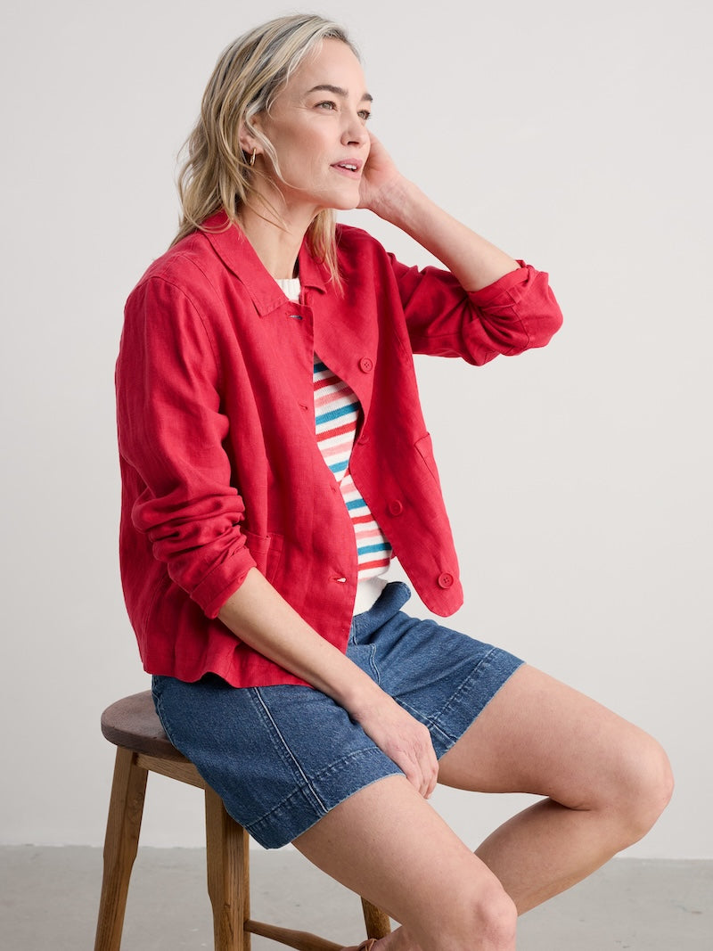 Woman wearing a pink jacket, striped shirt, and denim shorts sitting on a wooden stool against a plain background.