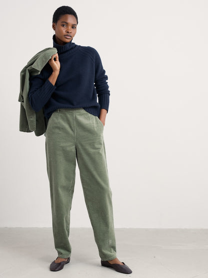 Person wearing a navy sweater and green pants holding a green jacket against a white background