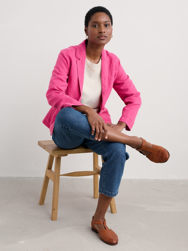 Woman wearing a bright pink blazer sitting on a wooden stool against a plain background