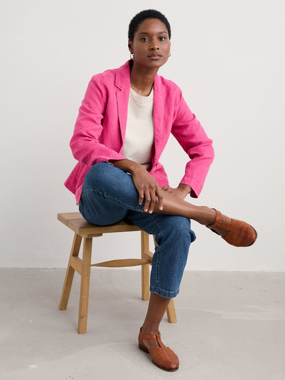 Woman wearing a bright pink blazer sitting on a wooden stool against a plain background