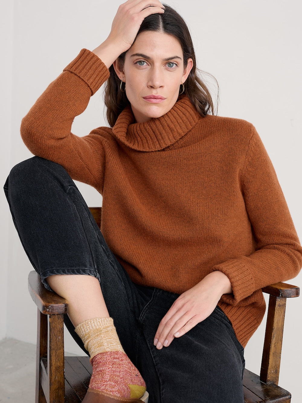 Woman wearing a brown turtleneck sweater sitting on a wooden chair.
