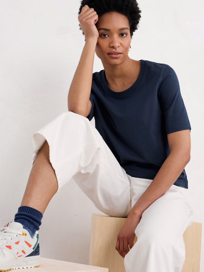 Woman wearing a navy t-shirt and white pants sitting on a chair against a plain background
