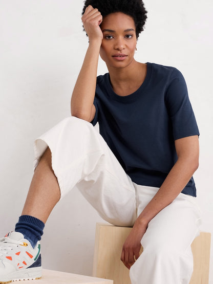 Woman wearing a navy t-shirt and white pants sitting on a chair against a plain background