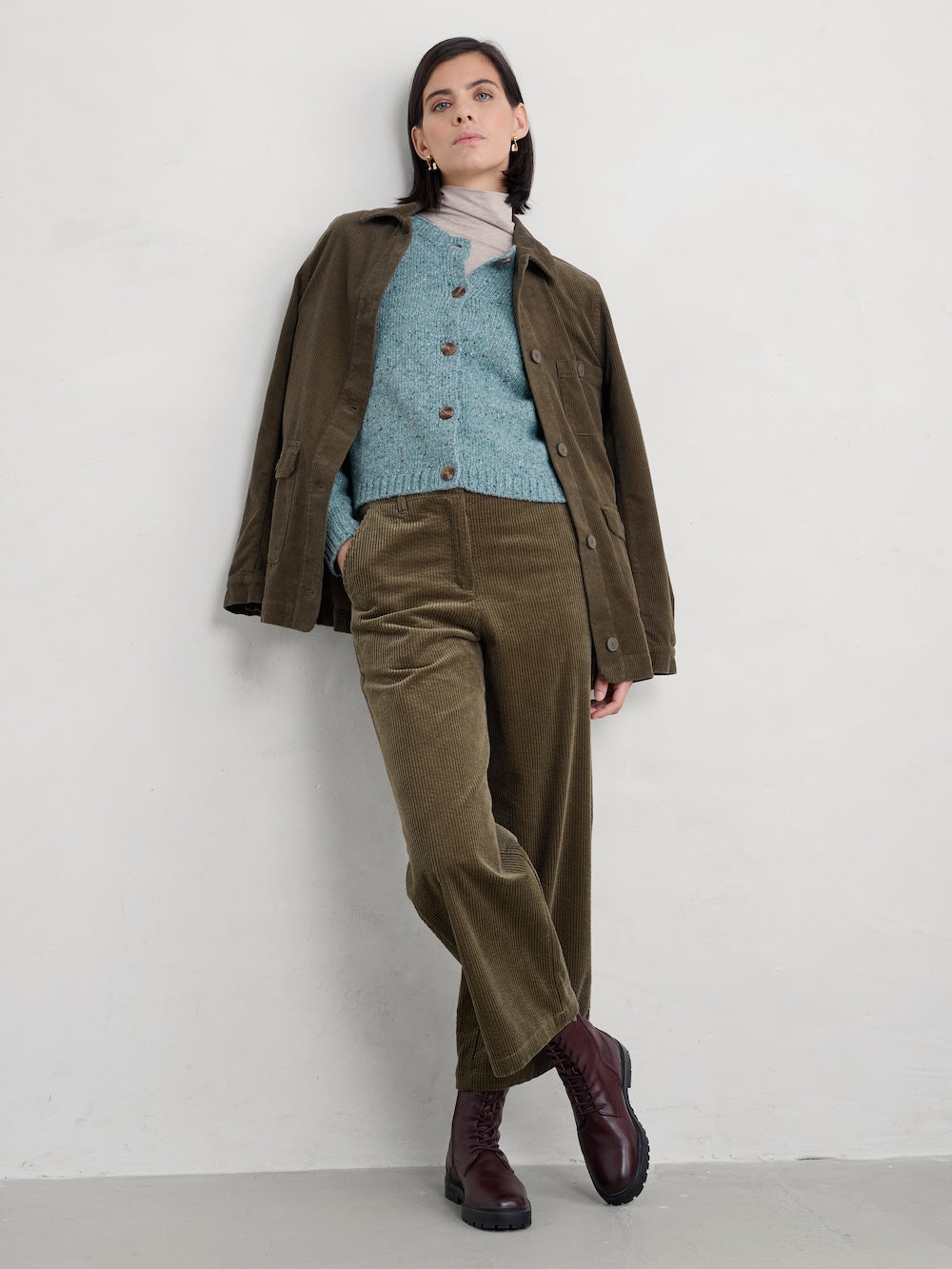 Woman wearing a brown jacket, blue sweater, and green pants against a white background