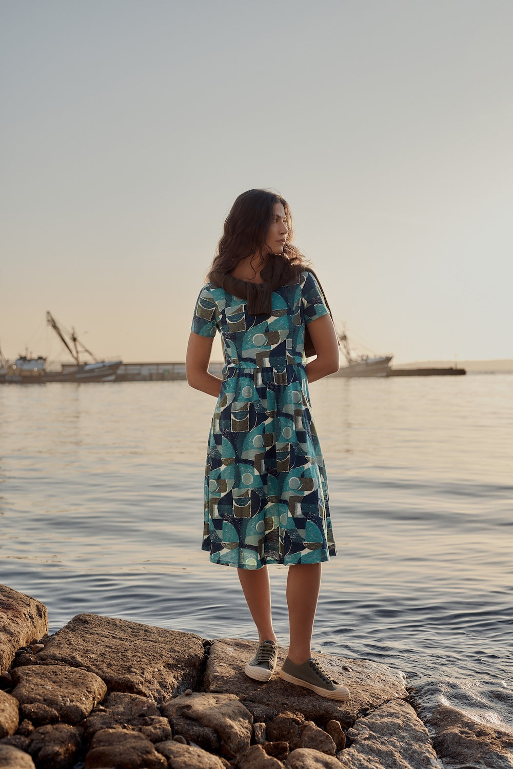 Woman in a patterned dress standing on rocks by a body of water.