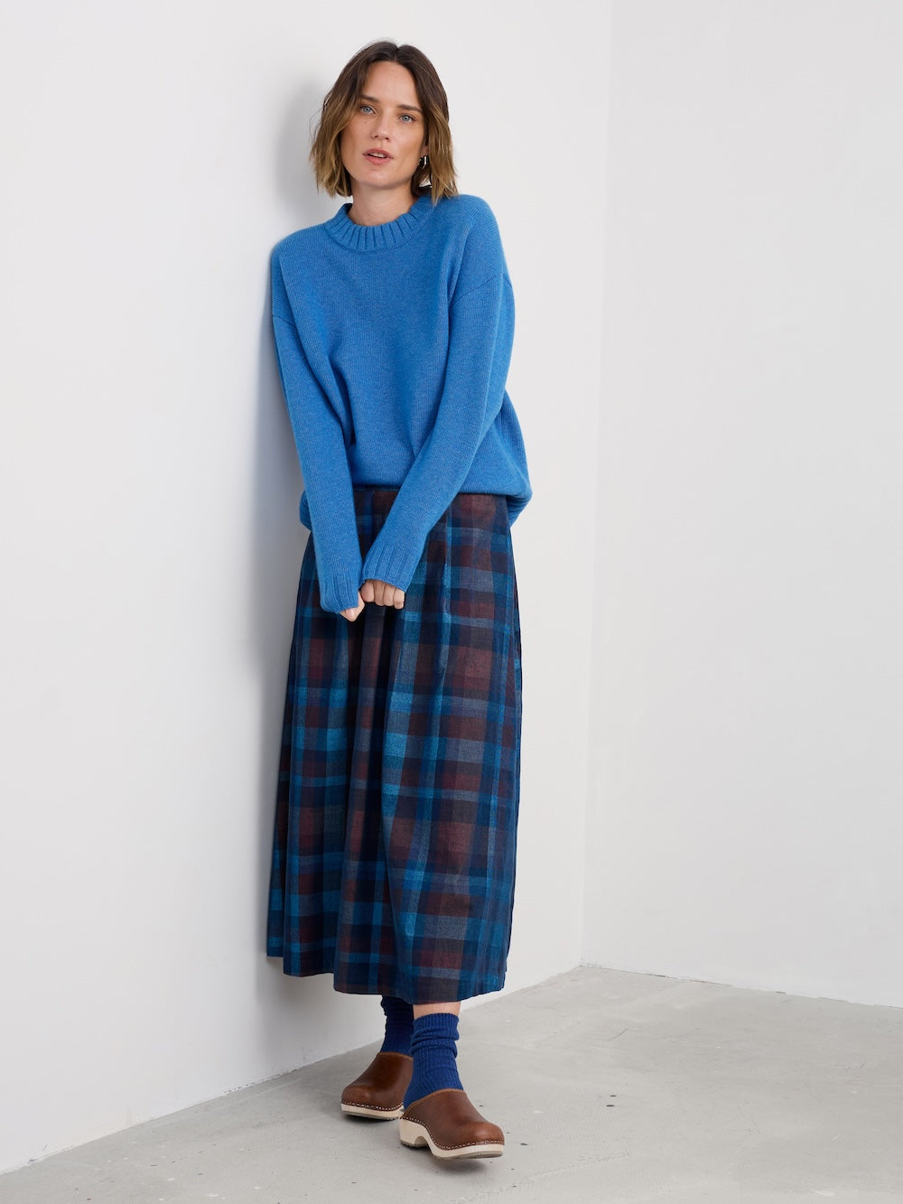 Woman wearing a blue sweater and plaid skirt against a white wall