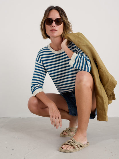Woman wearing a striped sweater and sunglasses, sitting on the floor with a jacket over her shoulder.