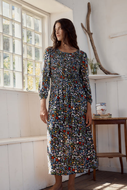 Woman wearing a floral dress standing in a room with a window and wooden shelf.