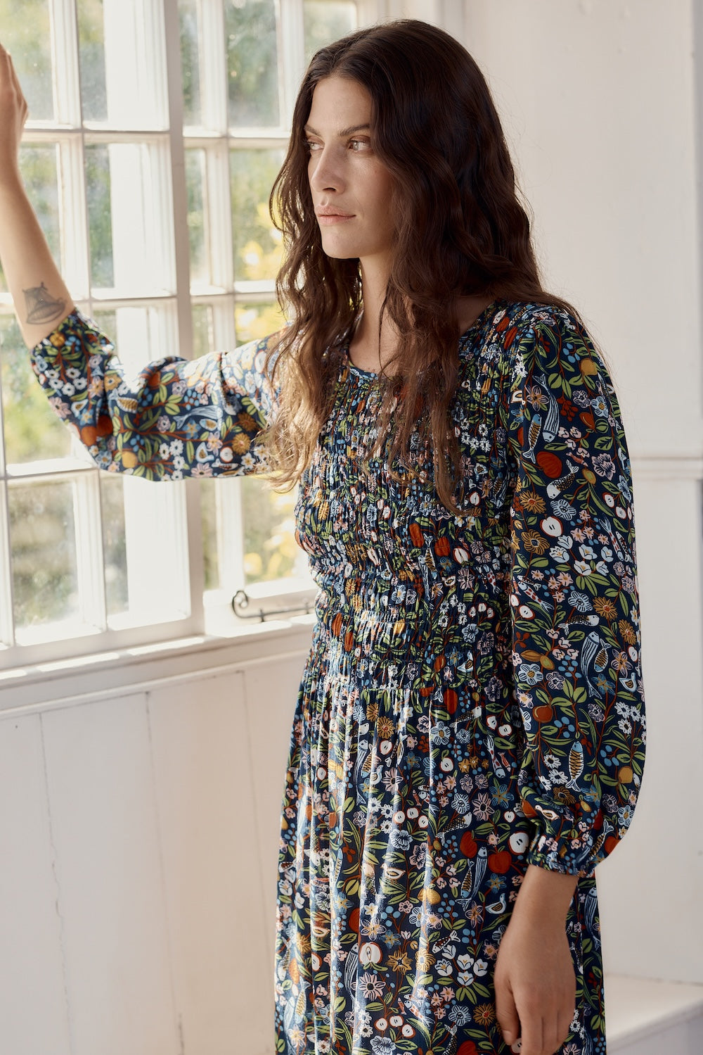 Woman wearing a floral dress standing by a window with white walls.