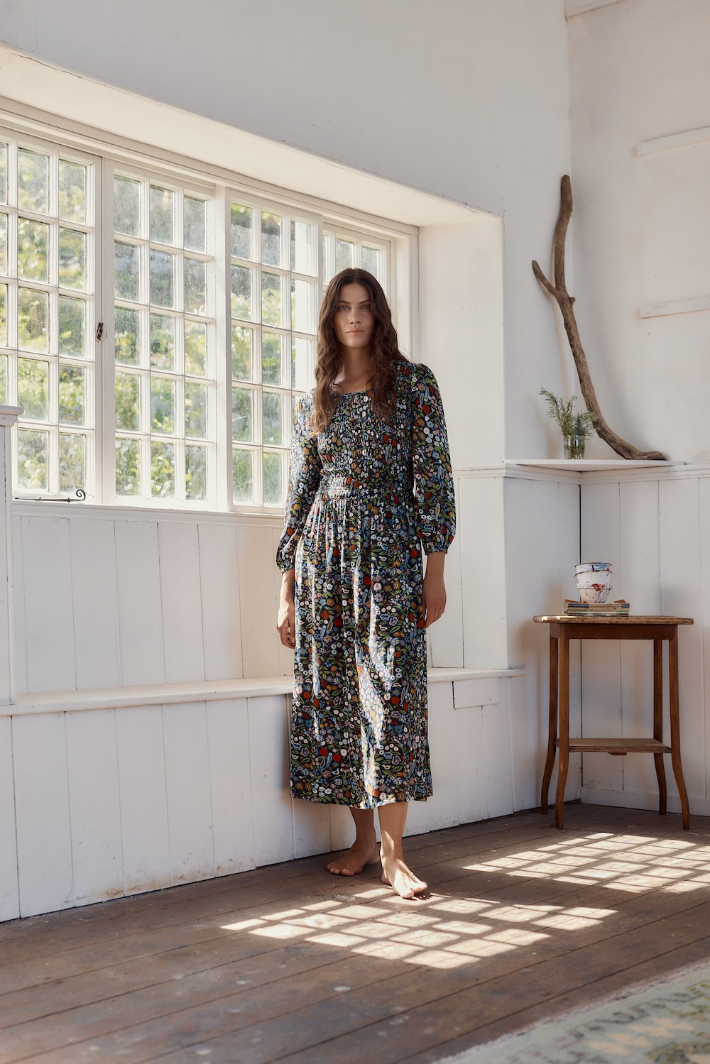 Woman in a floral dress standing in a sunlit room with wooden floors and white walls.