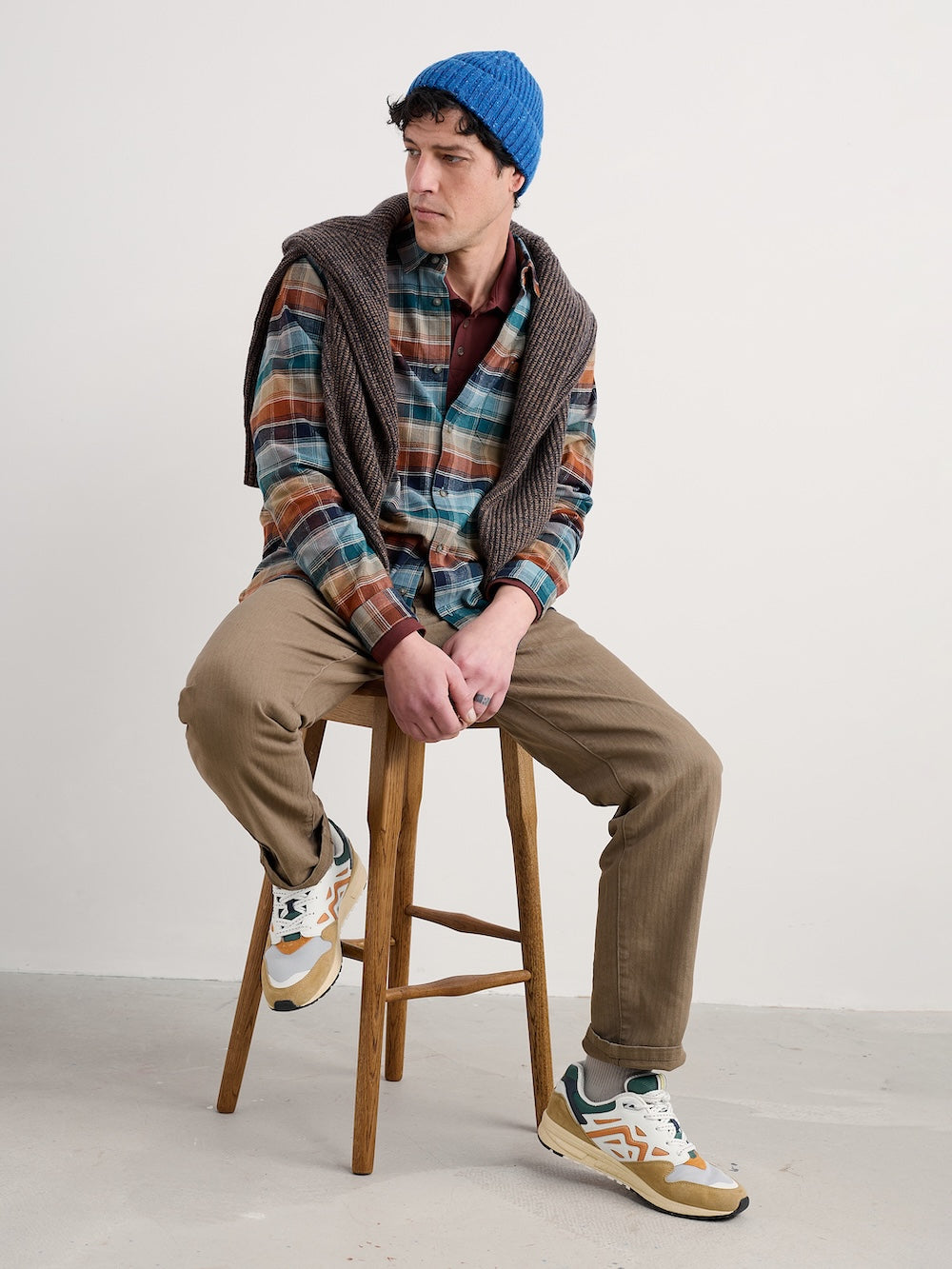 Man sitting on a stool wearing a plaid shirt, vest, and blue beanie against a plain background