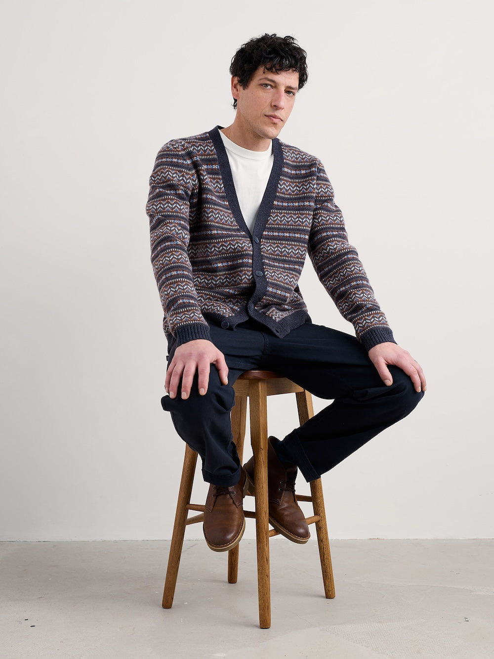 Man wearing a patterned cardigan sitting on a stool against a plain background