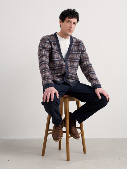 Man wearing a patterned cardigan sitting on a stool against a plain background