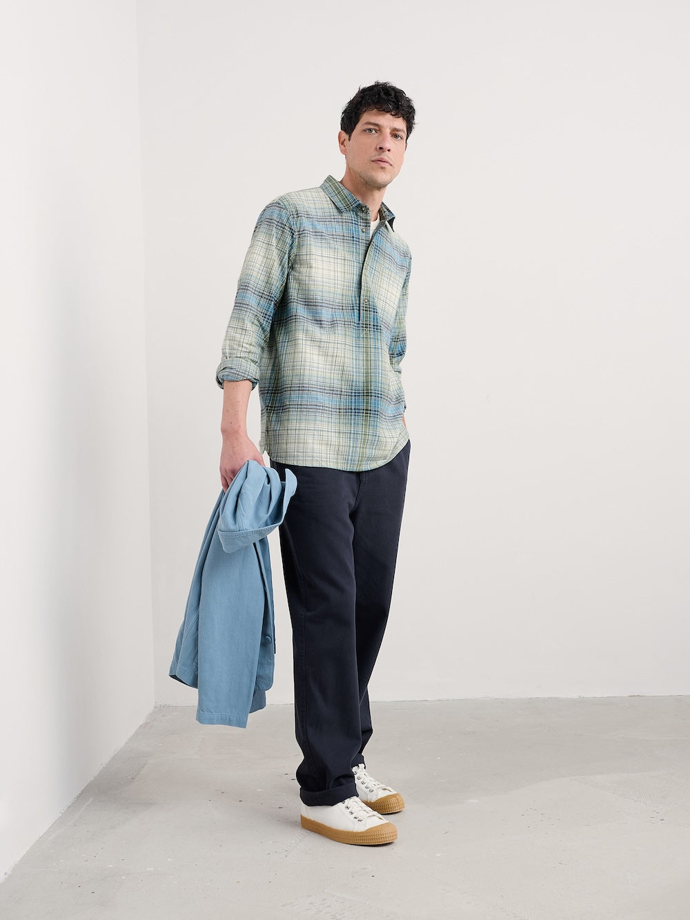 Man wearing a plaid shirt and dark pants holding a blue jacket against a white background
