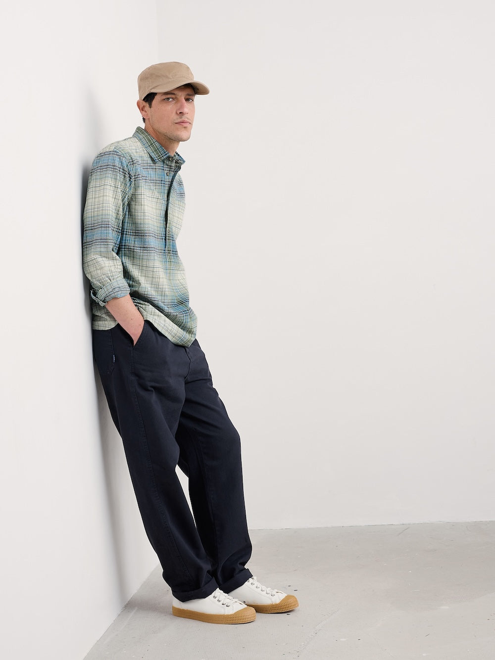 Man wearing a plaid shirt, cap, and dark pants against a white wall.