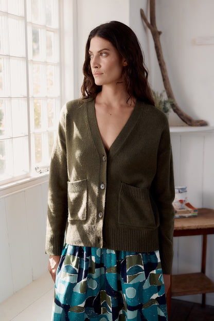 Woman wearing a green cardigan in a room with a window and wooden shelf.