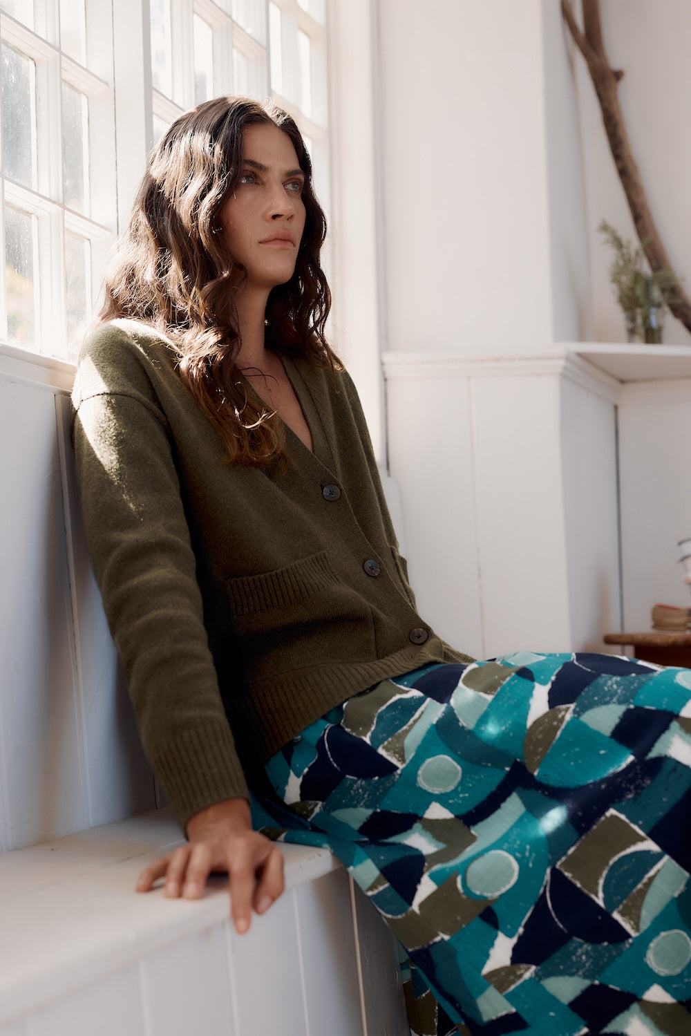 Woman wearing a green cardigan and patterned pants sitting by a window.