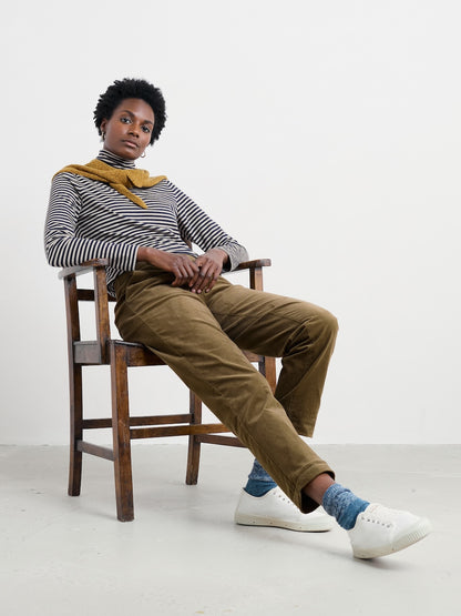 Person sitting on a wooden stool wearing a striped sweater, mustard scarf, brown pants, and white shoes.