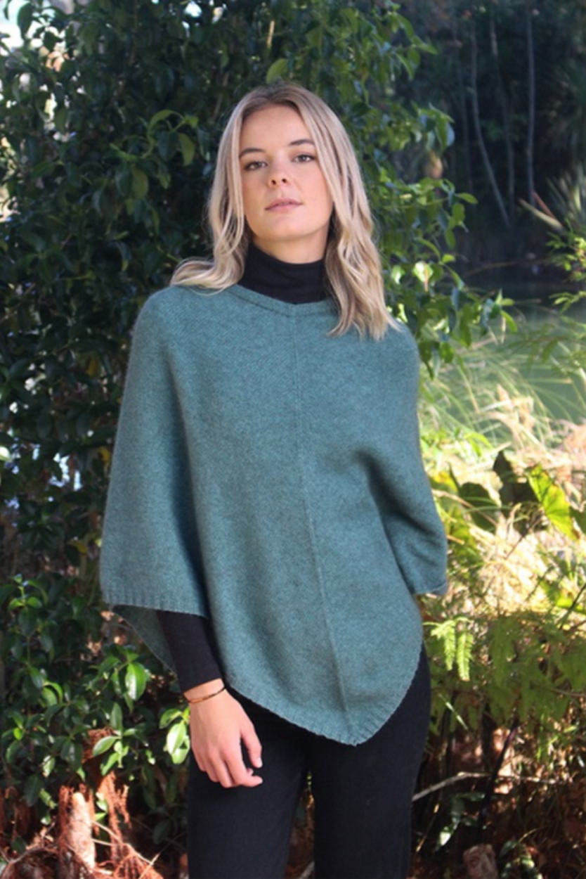 Sage green possum and merino wool poncho on female model standing in garden. Warm, soft, lightweight possum and merino wool. Lothlorian