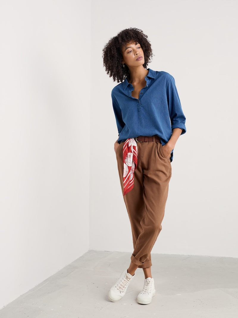Woman wearing a blue shirt and brown pants with a red and white pattern, standing against a white background.