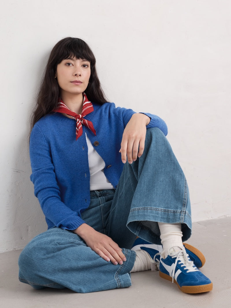 Person wearing a blue cardigan, white shirt, and jeans sitting on the floor against a white wall.