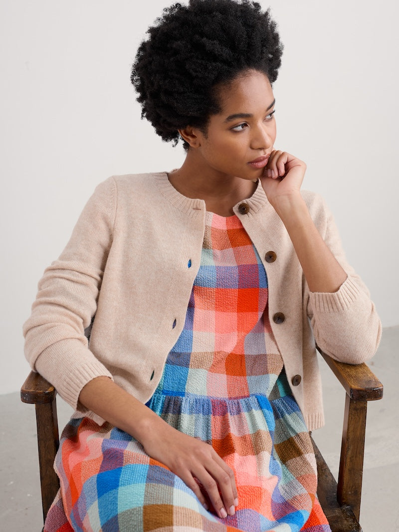 Woman wearing a beige cardigan over a colorful plaid dress, sitting on a chair.