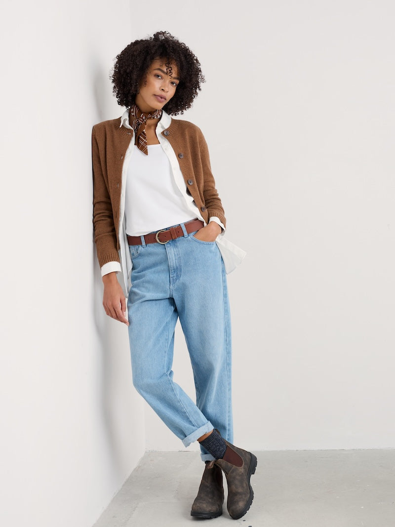 Woman wearing a brown cardigan, white shirt, blue jeans, and brown boots against a white background
