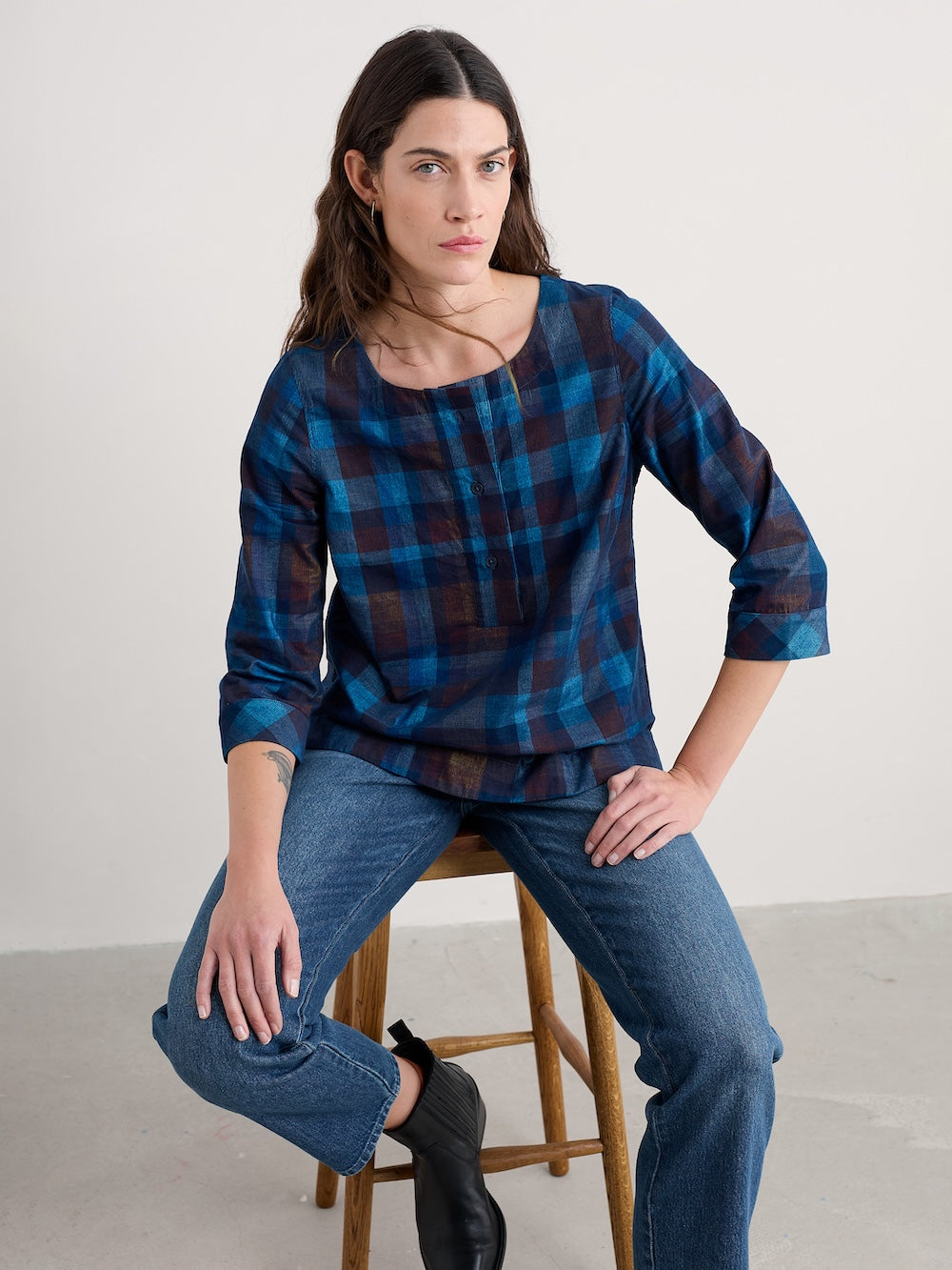 Woman wearing a blue plaid shirt and jeans sitting on a wooden stool against a white background