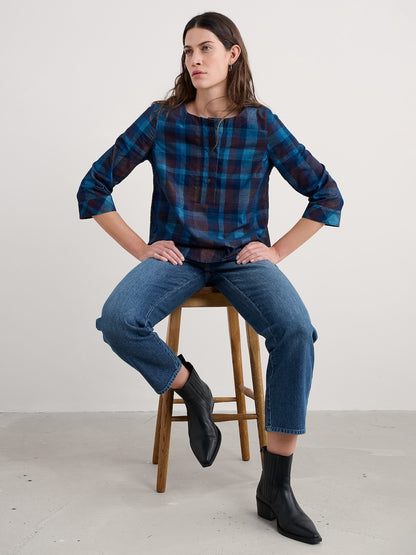 Woman sitting on a wooden stool wearing a blue plaid shirt and jeans.